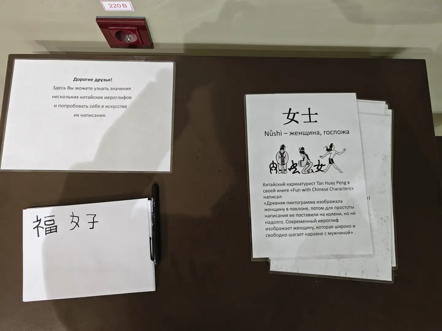 桌上放置着三张纸，其中一张写有“福”和“女子”两个汉字，旁边放着一支黑色笔。另一张纸上用俄语写着欢迎信息，邀请人们了解汉字并尝试书写。第三张纸上展示了一个汉字“女士”，并附有其拼音和俄语翻译，下方还有关于该字演变的解释和插图。