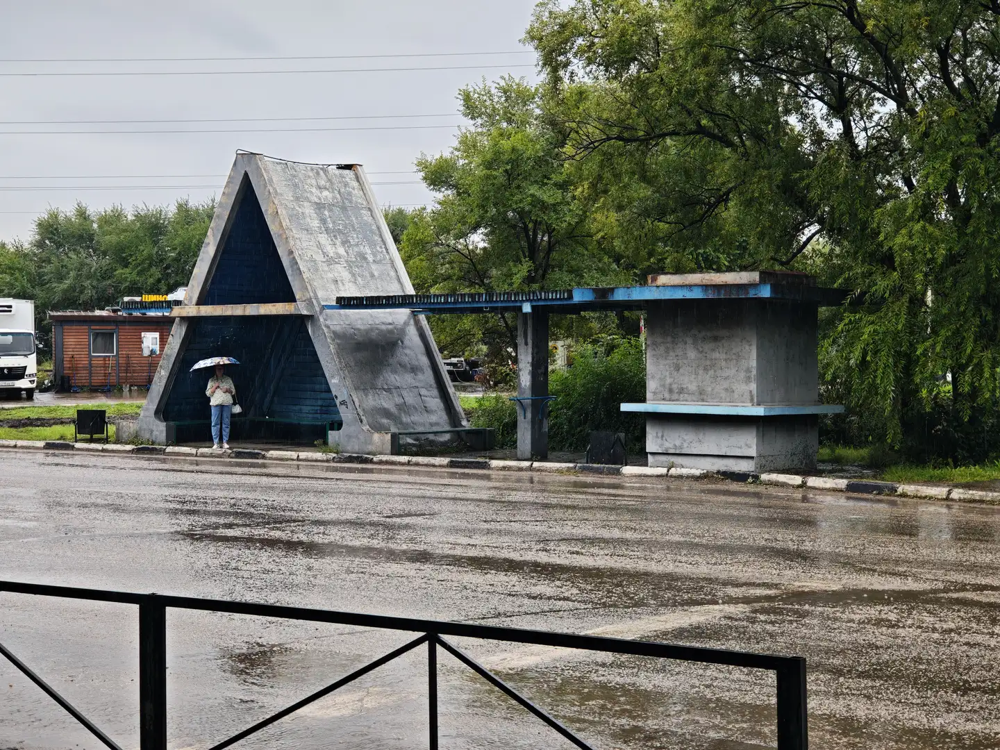 一个三角形结构的遮雨棚下站着一位撑伞的人，旁边是混凝土的长椅和蓝色顶棚。地面湿滑，有积水，背景中有树木和一辆白色货车。远处可见低矮的木屋和电线。