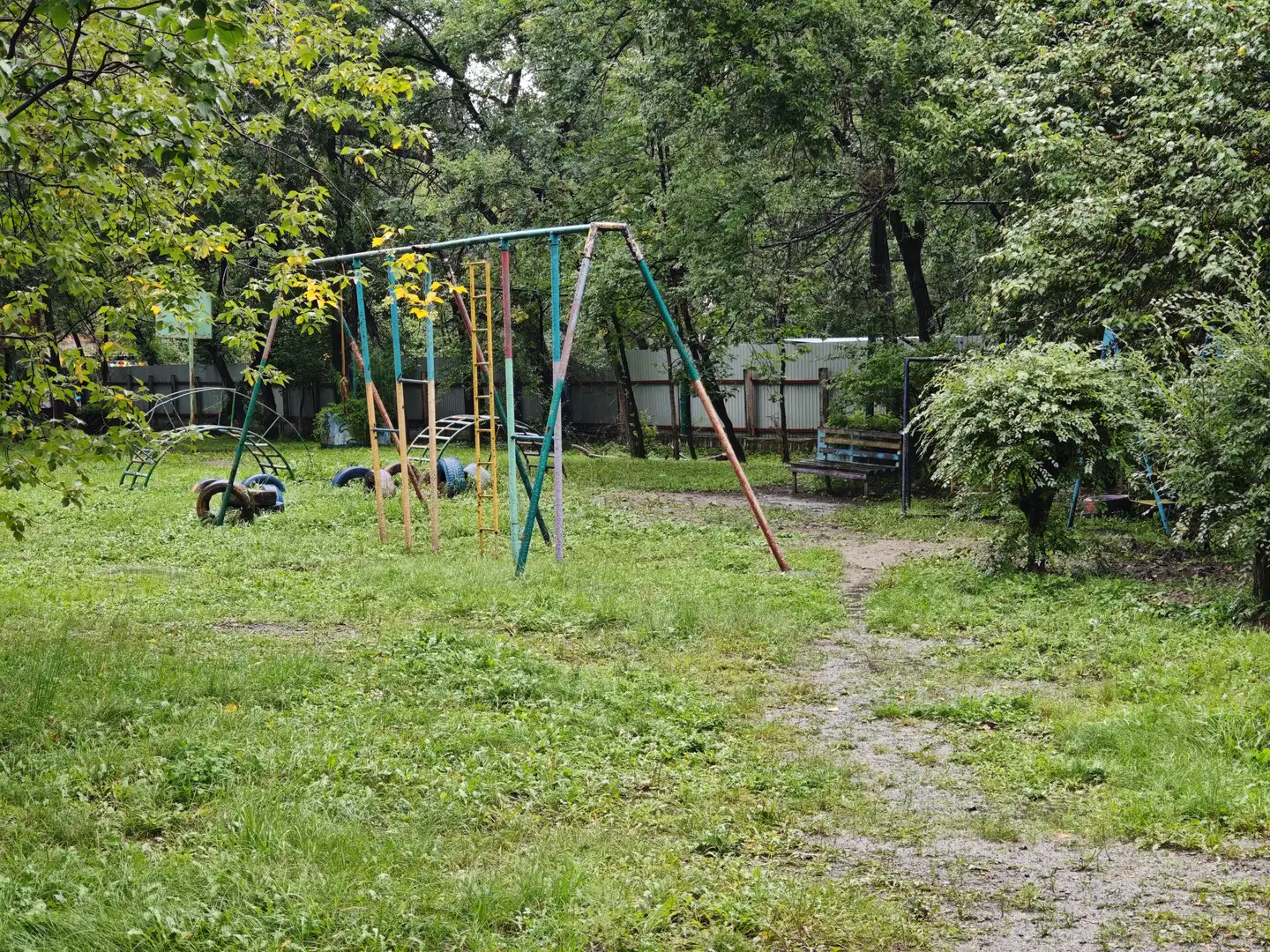 一个废弃的儿童游乐场，有生锈的秋千架和攀爬架，周围是茂密的草丛。一条小路穿过草地，通向远处的围栏和长椅。树木环绕，叶子开始泛黄。