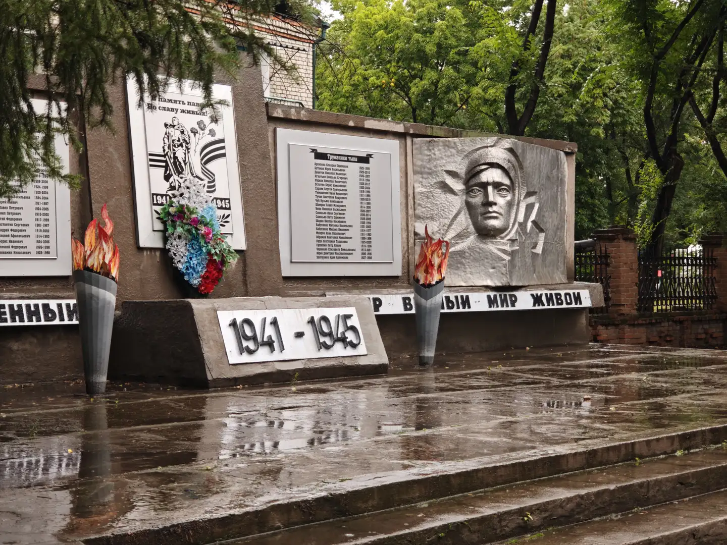 一座纪念墙上有浮雕和铭文，记录了1941至1945年的历史。两侧各有一个燃烧的火炬造型，前面是湿漉漉的台阶。墙上挂着花圈，背景是绿树和砖墙。