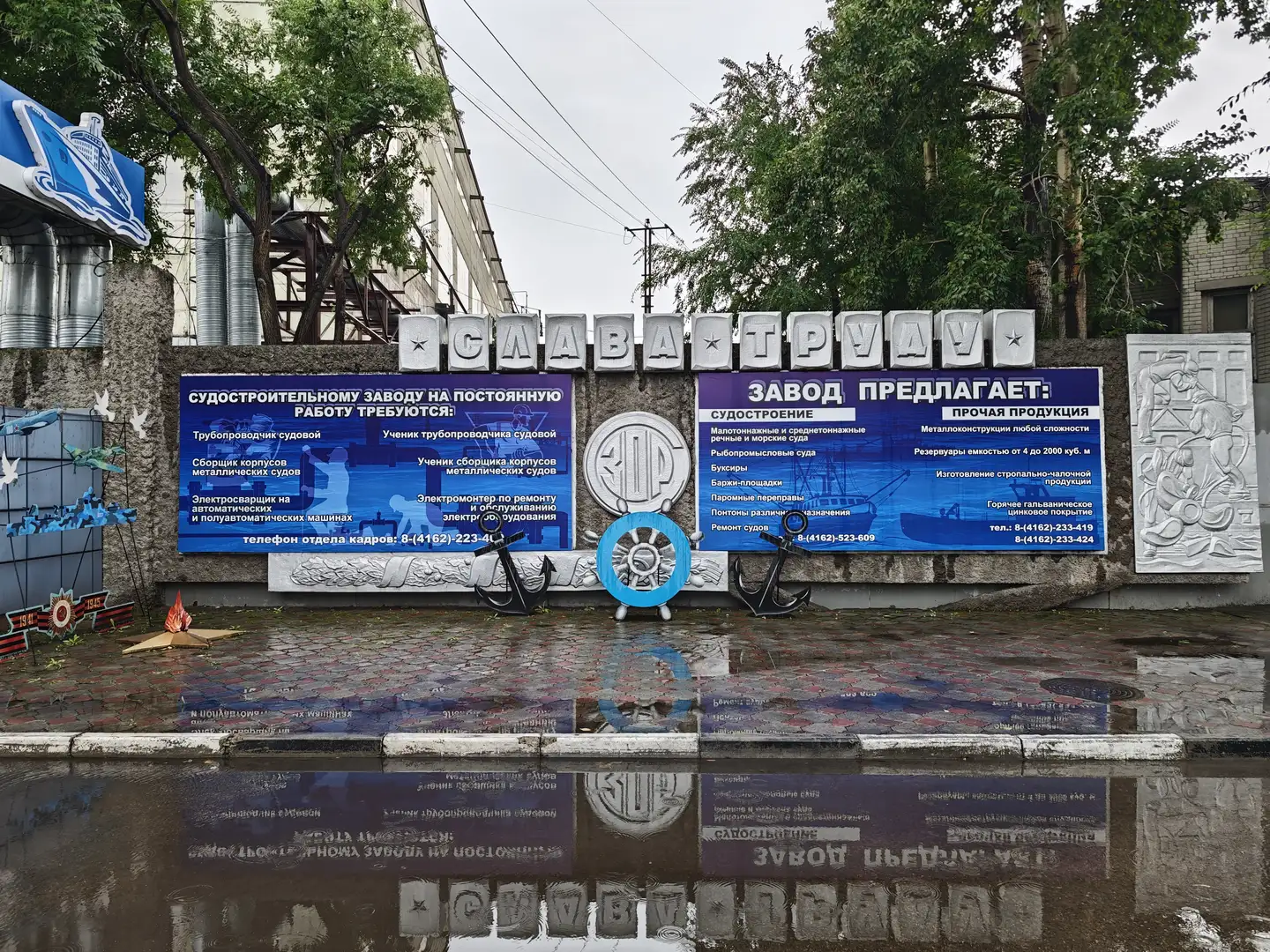 一面墙上写着“СЛАВА ТРУДУ”（劳动光荣），下方有招聘广告和船厂信息。墙前摆放着船锚和舵轮装饰，地面有积水反射文字。背景是工业建筑和电线杆。
