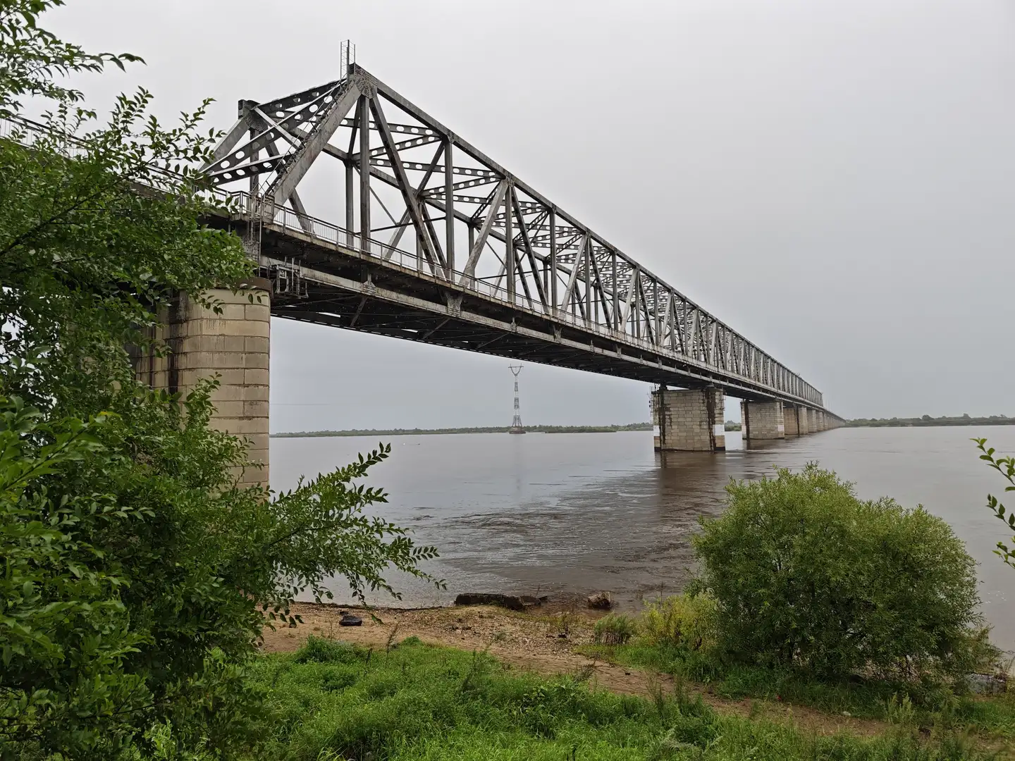 一座钢铁桁架桥横跨宽阔的河流，桥墩坚固，延伸至远方。两岸有绿色植被，河水呈浑浊棕色。天空灰蒙，天气阴沉。