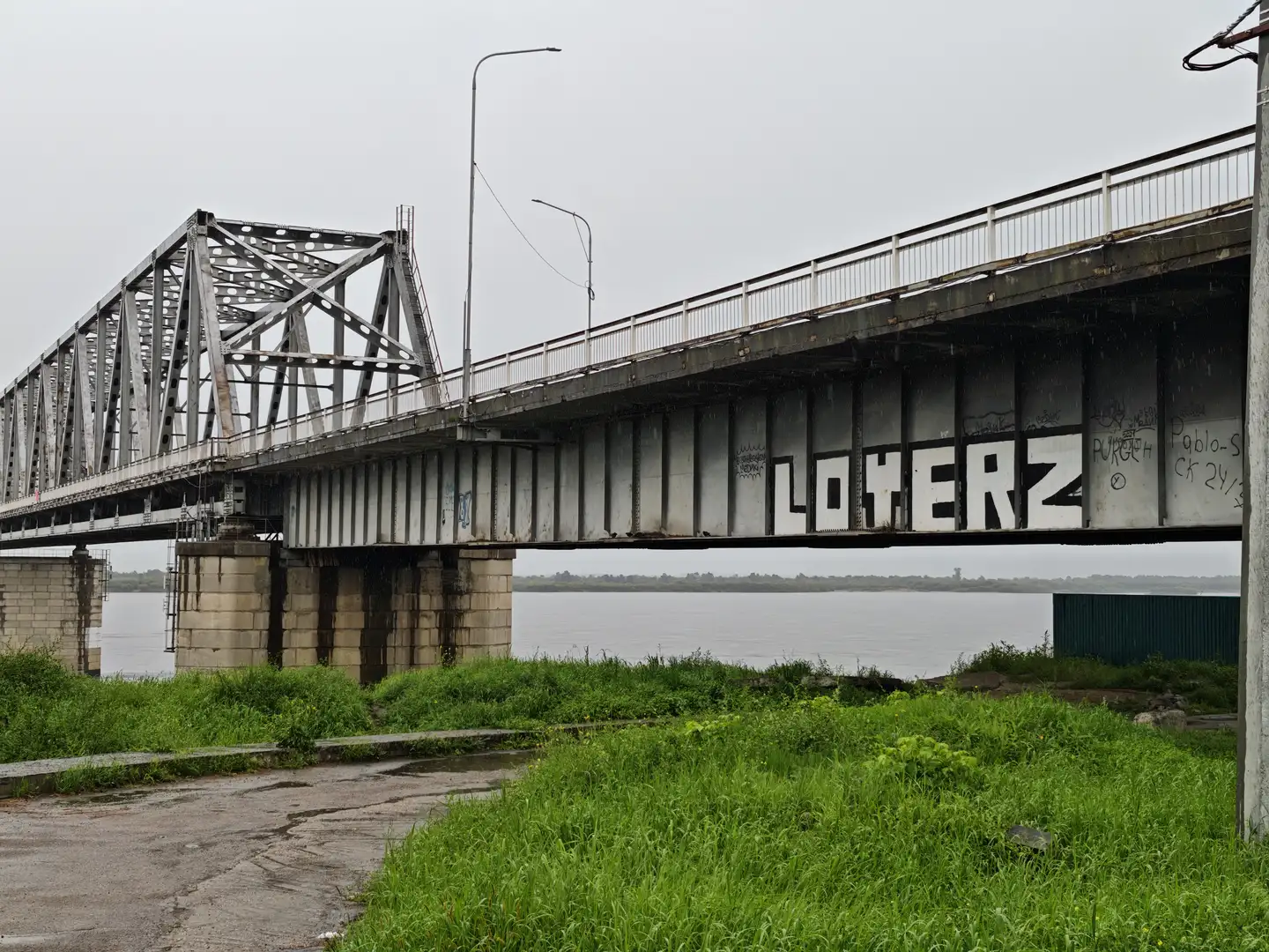 一座桥梁的侧面涂有“LOHERZ”字样，桥下是绿色的草地和一条小路。桥体由金属构成，支撑柱为混凝土。远处是宽阔的水面和对岸的景物。