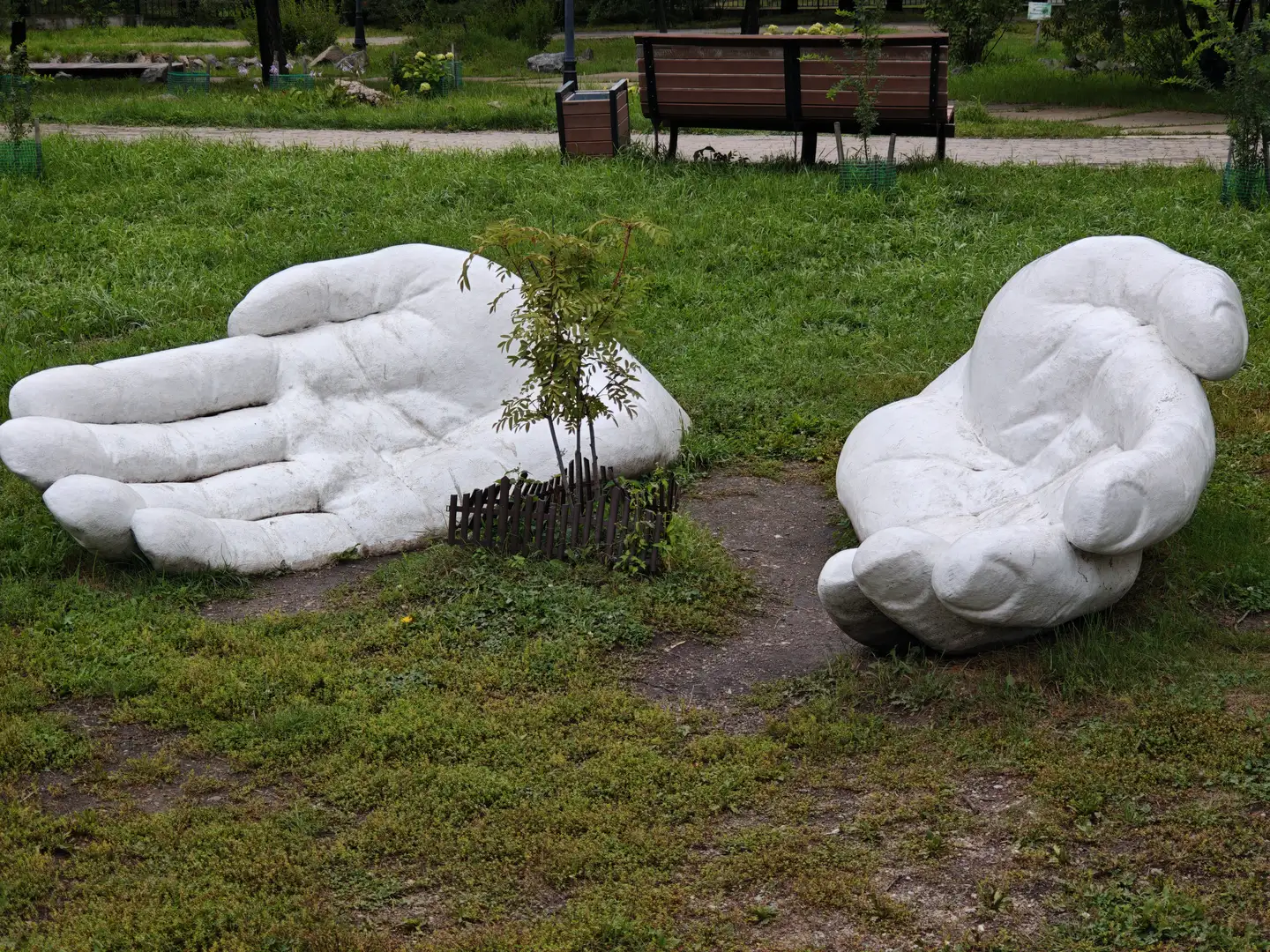 两只巨大的白色手掌雕塑并排置于草地上，其中一只手掌中有一棵小树被木栅栏围住。周围是绿色的草地和一条小路，远处有长椅和垃圾桶。背景中可以看到一些灌木和树木。