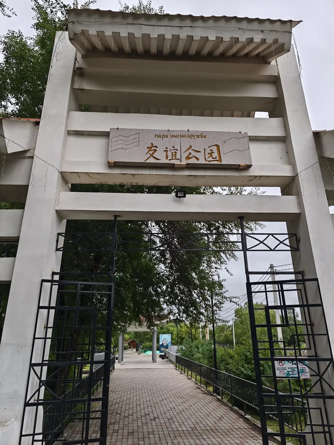 一座白色的拱门式入口，上方悬挂着写有“友谊公园”字样的标牌，标牌上有俄文和中文文字。入口由黑色金属栅栏构成，通往一条铺有砖块的小路。小路两侧有绿树和电线杆，远处可见一个蓝色广告牌。