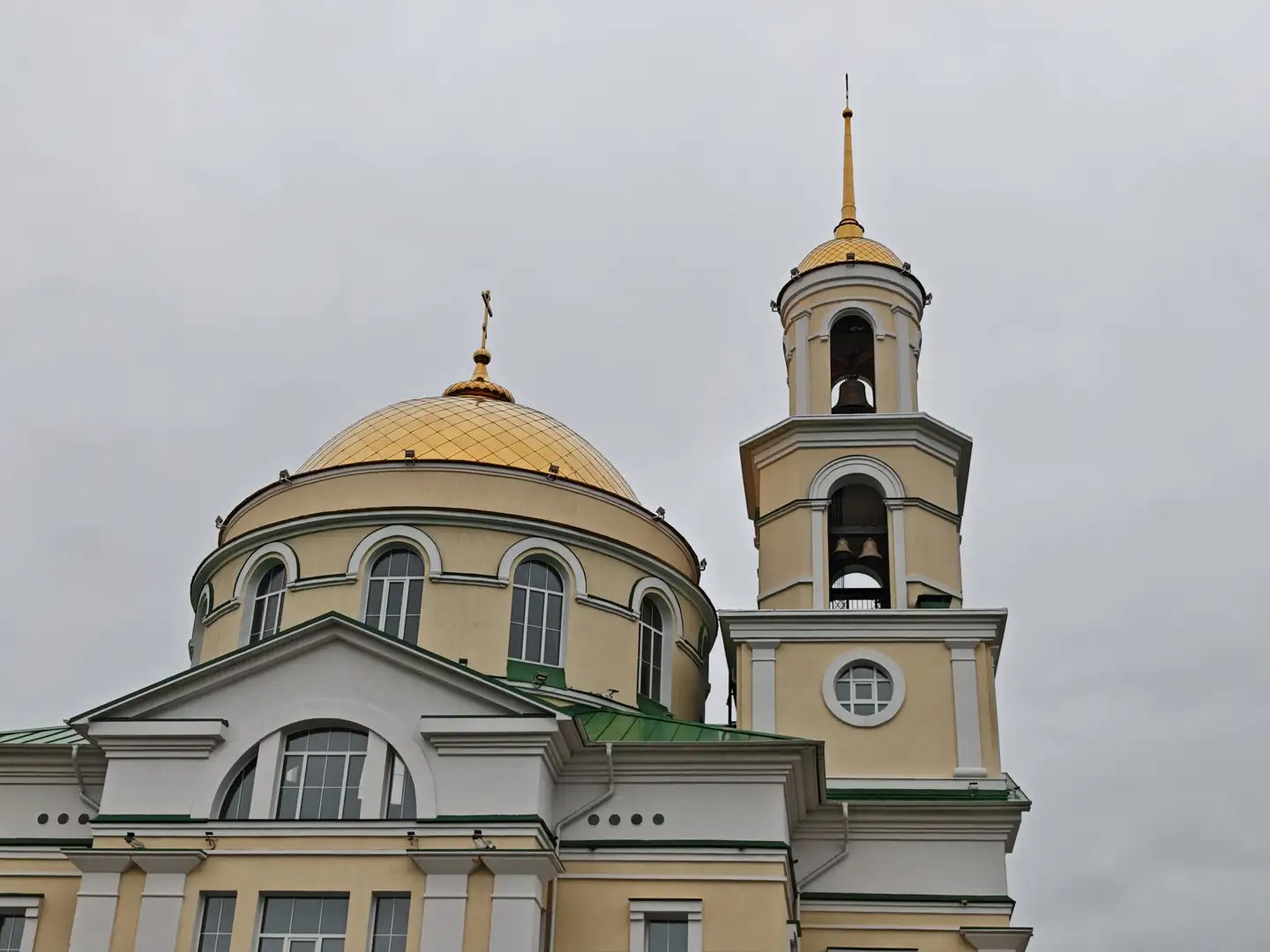 一座黄色和白色相间的教堂建筑，顶部有一个金色圆顶和一个高耸的钟楼，钟楼顶端是金色尖顶。圆顶上有一个十字架，钟楼内可见悬挂的钟。建筑正面有多扇拱形窗户，屋顶边缘为绿色。