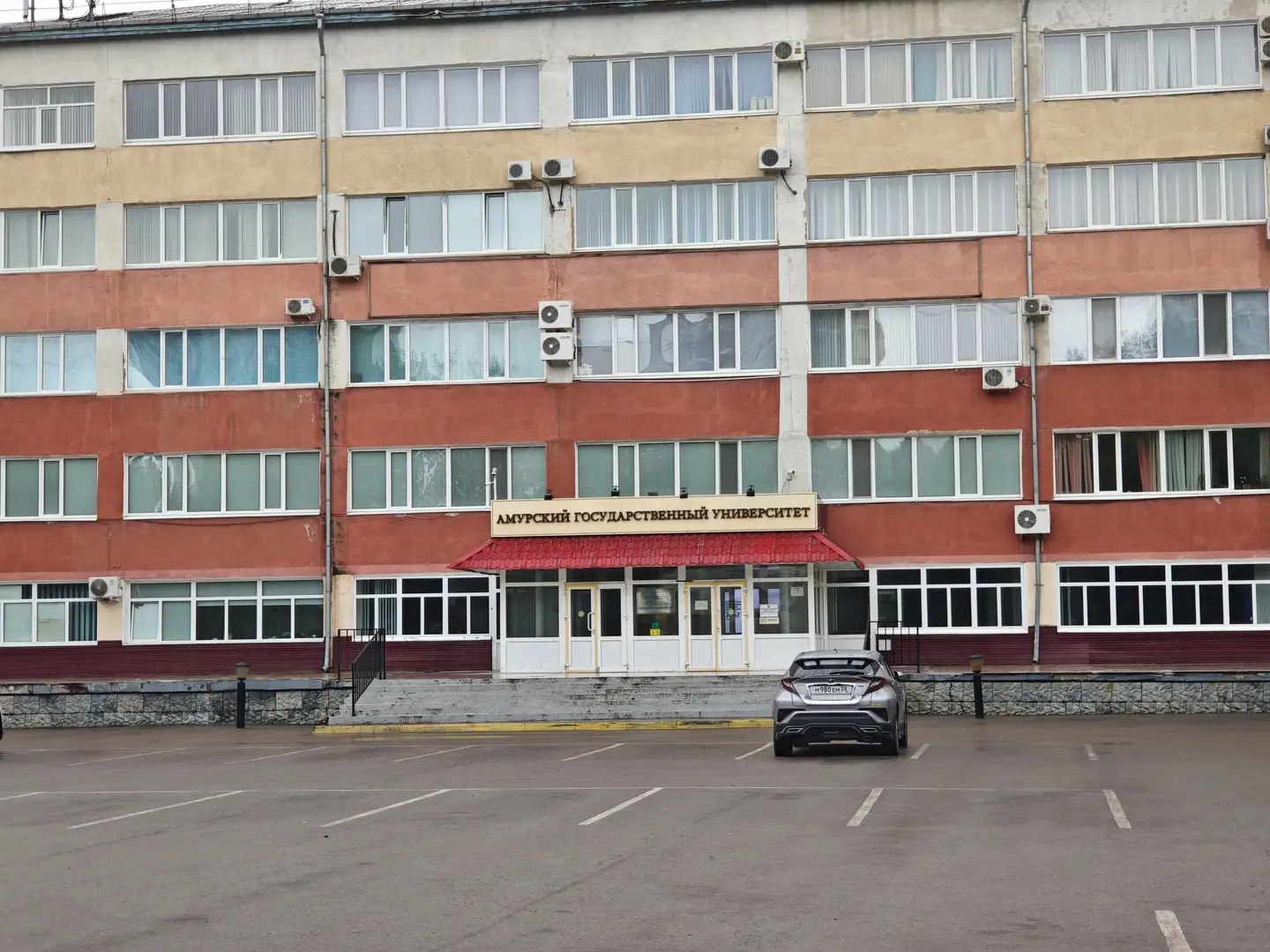 一栋多层建筑，外立面为红黄相间，窗户排列整齐，墙上安装多个空调外机。建筑正门上方有一个红色遮阳棚，上面写着“阿穆尔国立大学”的俄文名称。门前停着一辆银色汽车，地面为灰色停车场。