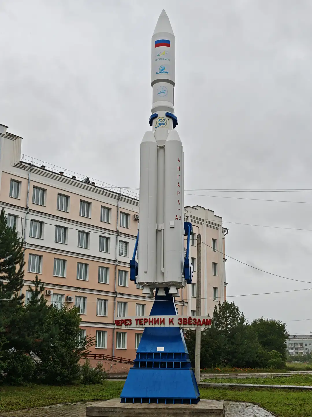 一座大型火箭模型矗立在蓝色底座上，火箭主体为白色，带有蓝色和红色装饰，侧面写有“安加拉-A5”字样。火箭底部有红色标语，写着“通过荆棘走向星辰”。背景是一栋浅色多层建筑，周围有绿树和草地。