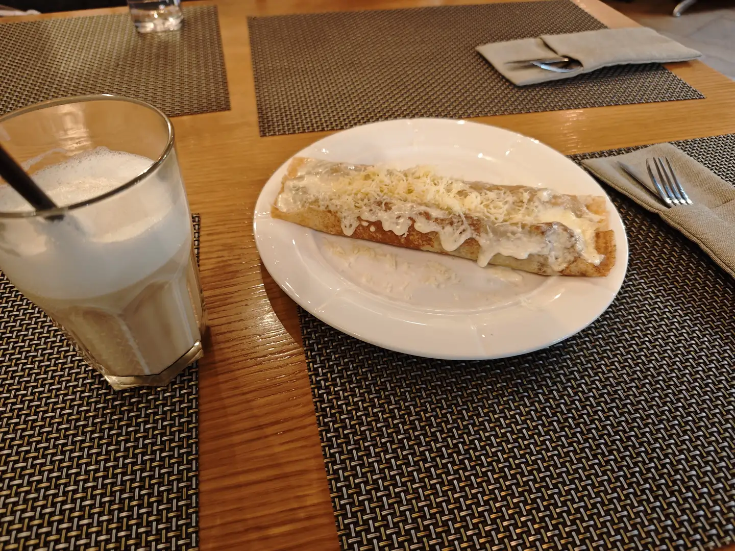 一张白色餐盘上放着一份卷起的煎饼，上面覆盖着奶油和碎奶酪，旁边是一杯装有泡沫饮料的透明玻璃杯。餐盘放在深色编织餐垫上，桌面上还有其他餐具和餐垫。背景中可见木质桌面和另一套餐具。