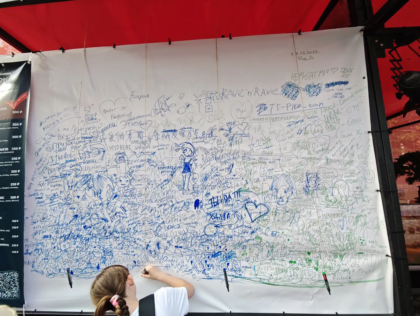 一位女孩正在一块大白布上用笔写字，白布上已经写满了各种颜色的涂鸦、签名和图画。白布悬挂在红色帐篷下，左侧有一个黑色广告牌，右侧有几支笔插在布上。背景中可见部分人群和树木。