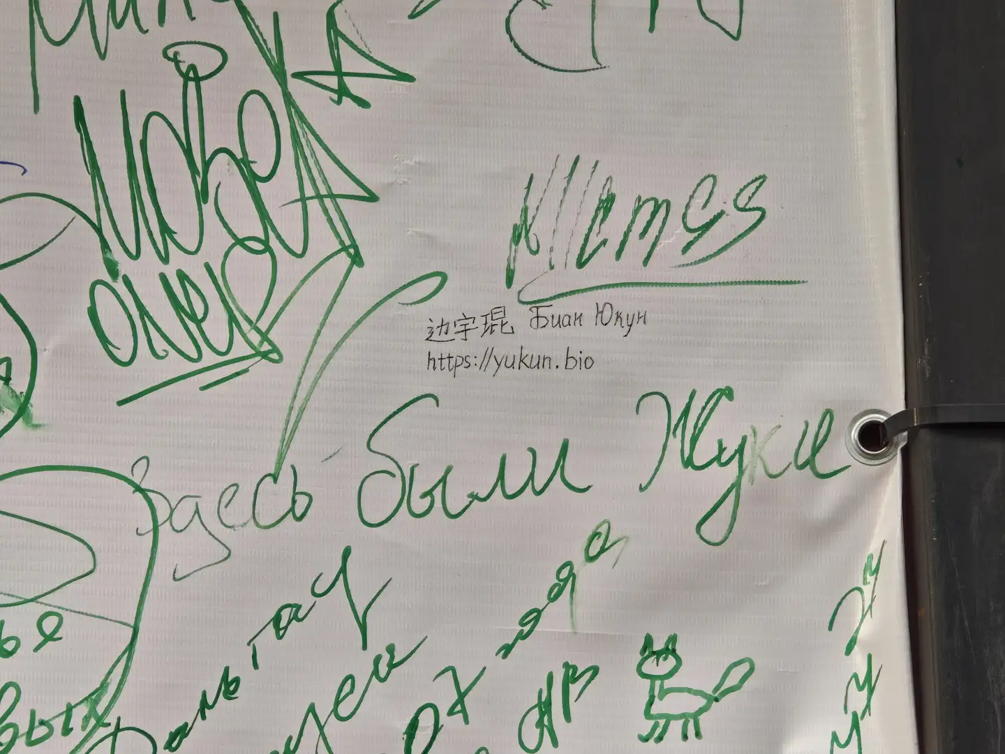 白布上用黑色笔迹写着“边宇琨 Bian Yukun https://yukun.bio”，旁边有其他涂鸦和文字。布料边缘有金属扣眼，右侧可见黑色框架。布上还有其他手写内容和简笔画。