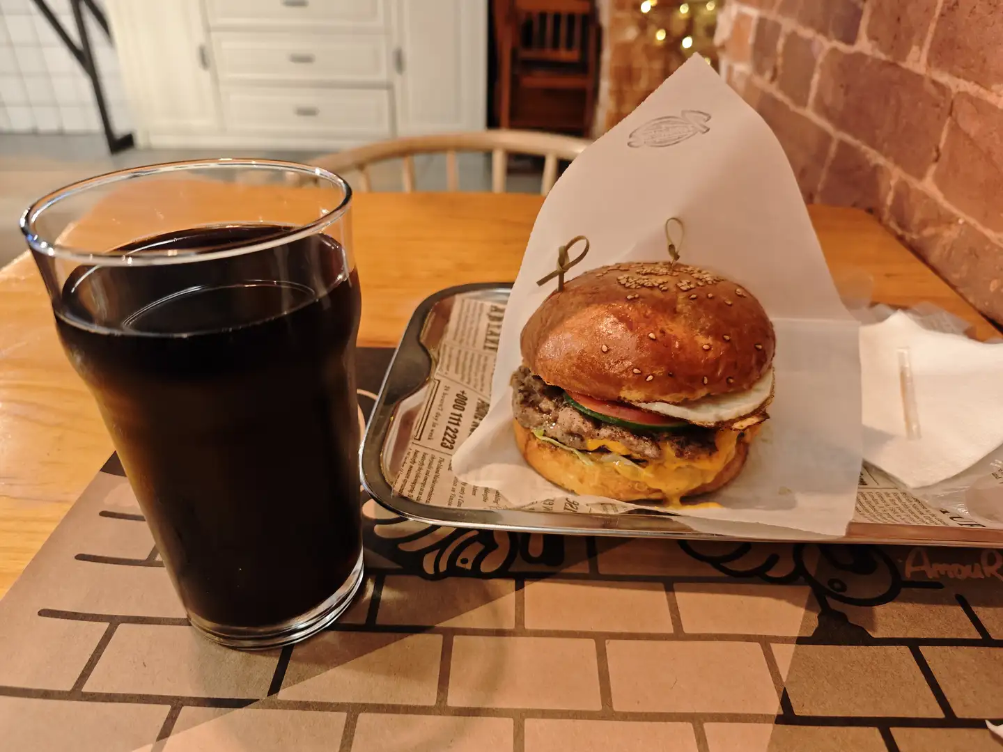 一个汉堡放在白色纸托上，置于金属托盘中，汉堡由芝麻面包、肉饼、生菜、番茄和芝士组成。旁边是一杯深色饮料，放在木质桌面上。背景是砖墙和白色柜子，桌上铺有印有图案的餐垫。