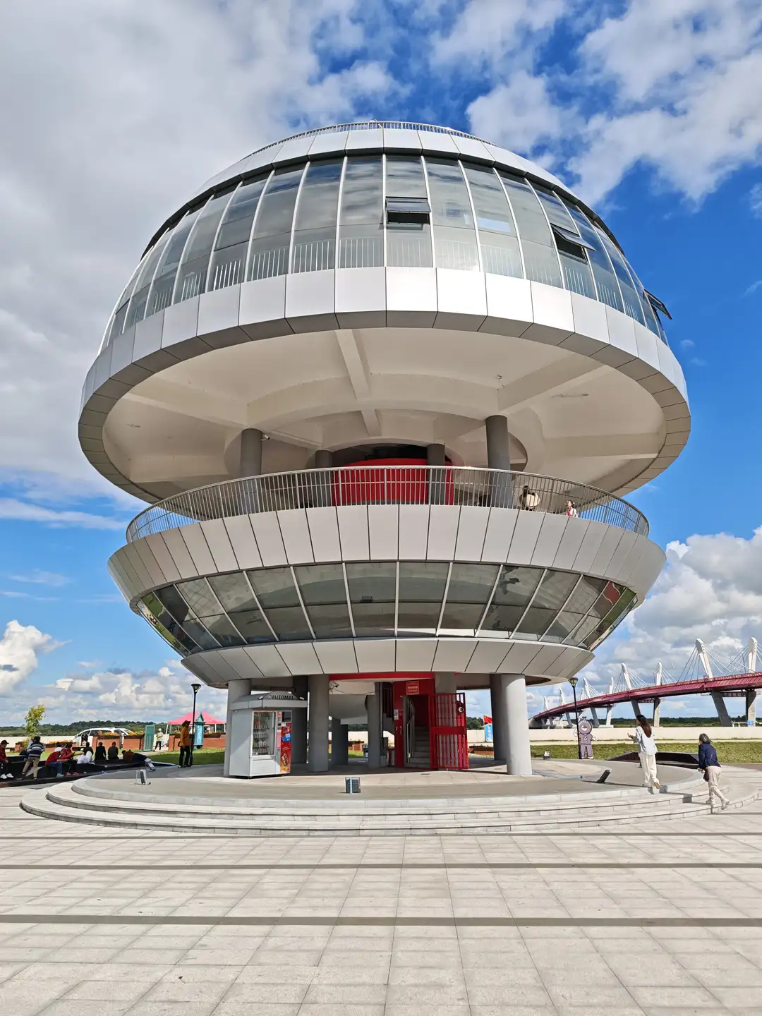 一个圆形的现代建筑，顶部有玻璃幕墙和金属栏杆，下方有红色的门和自动售货机。建筑周围是开阔的广场，地面铺有灰色地砖，远处可见一座红色桥梁。天空晴朗，有少量白云。
