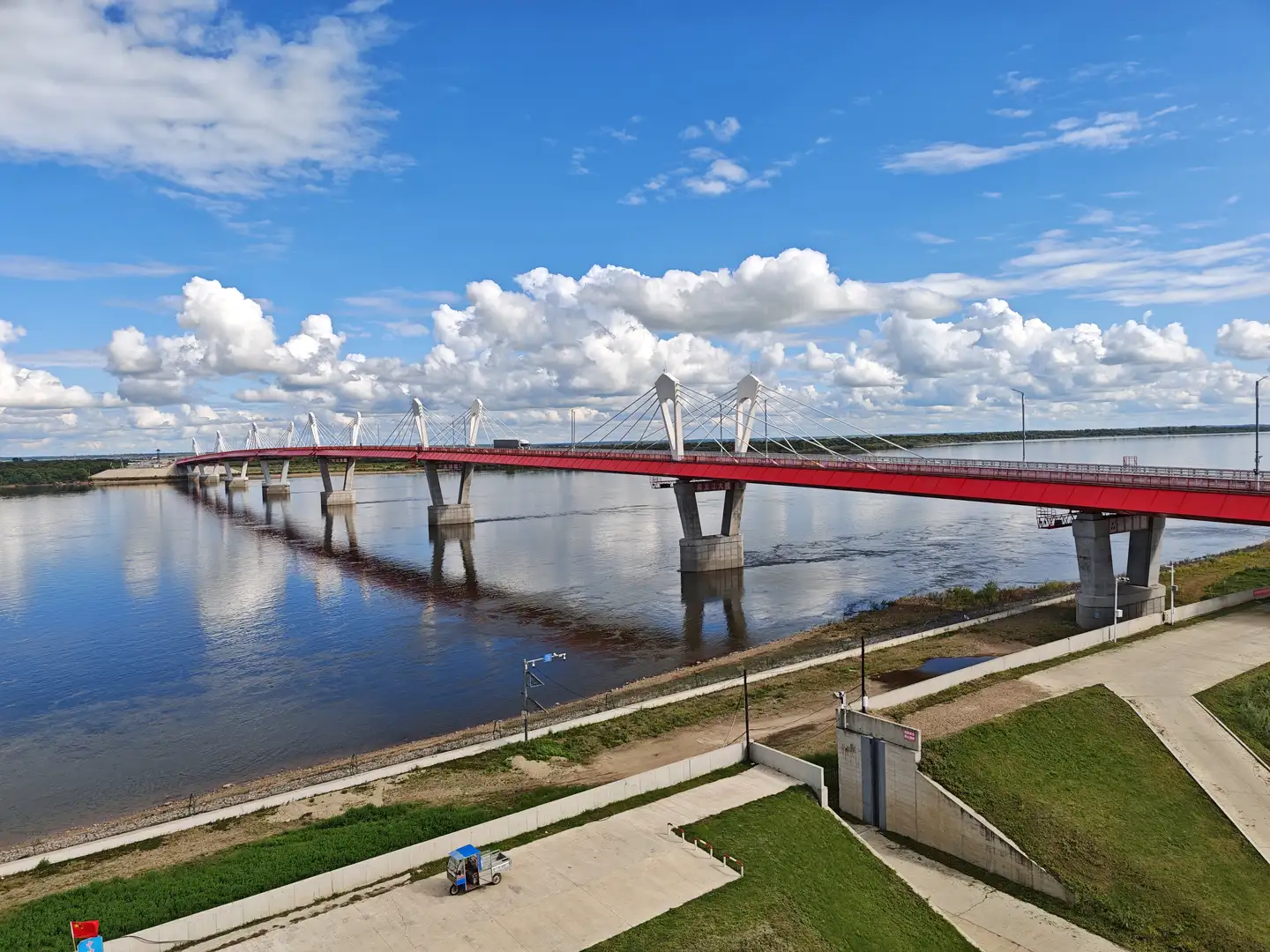 一条宽阔的河流上横跨着一座红色的斜拉桥，桥身由多个混凝土桥墩支撑。河岸两侧有绿化带和小路，岸边停着一辆蓝色顶棚的小车。天空湛蓝，布满蓬松的白色云朵。