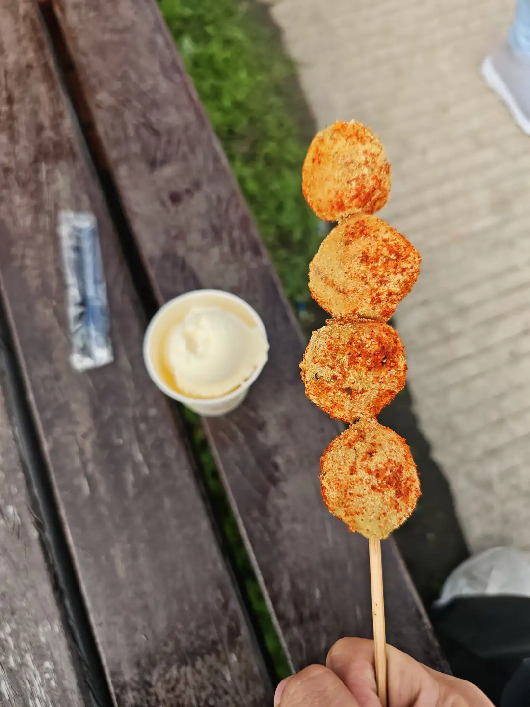 一只手拿着一串炸得金黄的丸子，丸子表面撒有红色调料。旁边是一杯白色的酱料，放在木制长椅上。背景是户外的草地和人行道。