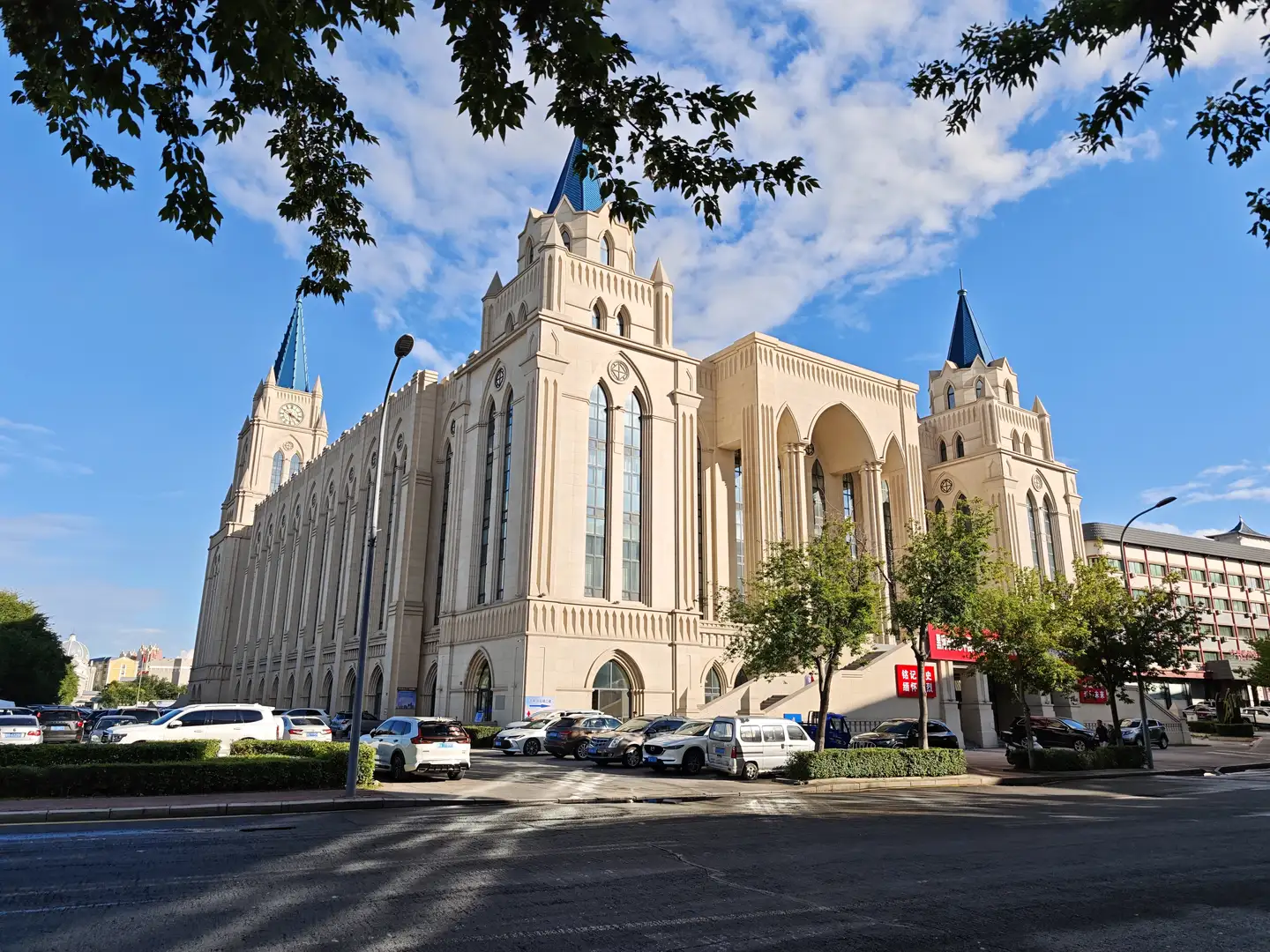 一座大型的欧式风格建筑，米黄色墙体，带有尖顶塔楼和拱形窗户。建筑前有停车场，停有多辆汽车，周围种有绿树。天空晴朗，有几片白云。