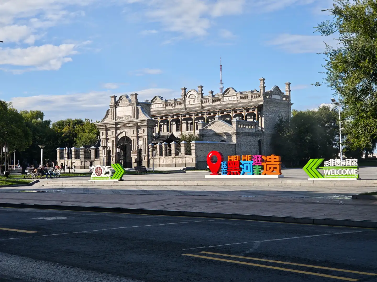 一座具有历史感的石砌建筑，正面有拱形门洞和装饰性柱子，门前有“我在黑河非遗”的彩色标识。建筑前方是宽阔的广场，周围有树木和行人。天空晴朗，阳光明媚。