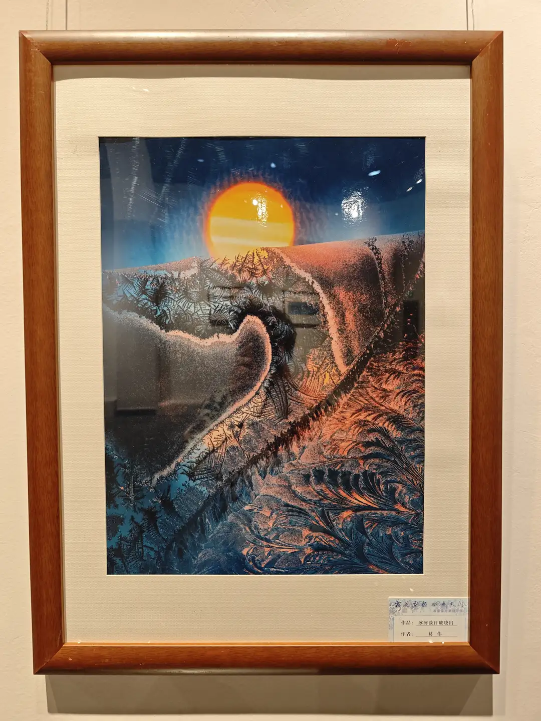 一幅装裱在木质画框中的艺术作品，描绘了日出或日落时分的山景，画面中有橙色的太阳和深蓝色的天空。山体轮廓用黑色线条勾勒，呈现出独特的纹理效果。画作下方有中文标签，标明作者和作品名称。