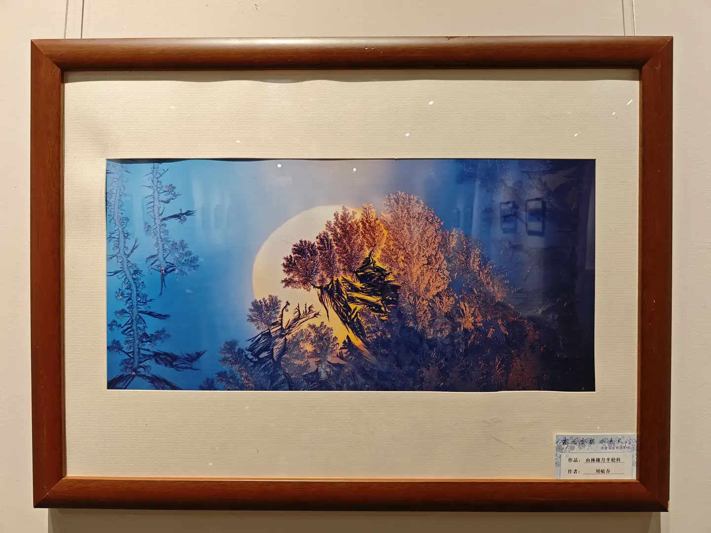 一幅装裱在木质画框中的艺术作品，描绘了月光下的山林景色，月亮呈淡黄色，周围是深蓝色的夜空和橙色的树木。画面左侧有几棵细长的树影，整体构图宁静而神秘。画作下方有中文标签，标明作者和作品名称。