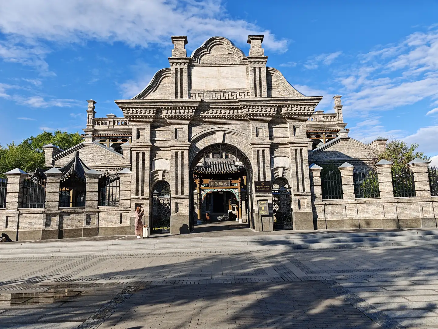 一座中式传统风格的建筑大门，由灰色砖石砌成，门楣上有精美的雕刻和装饰。大门上方悬挂着一块牌匾，门前有台阶和护栏。天空晴朗，有几片白云。