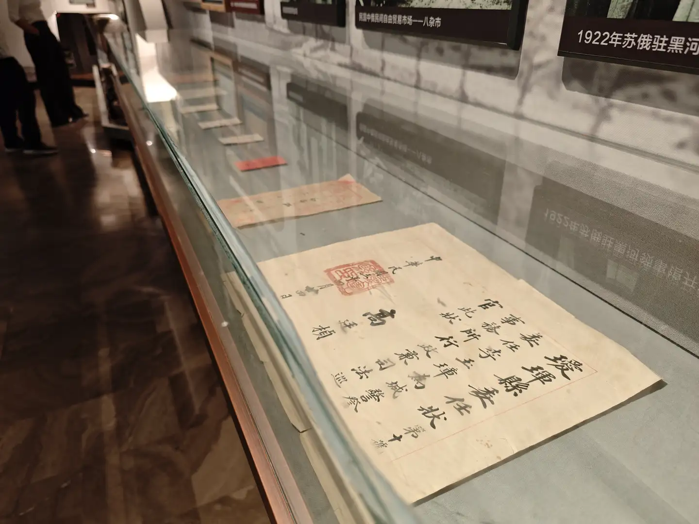 一个玻璃展柜内陈列着几张泛黄的旧纸张，上面有毛笔书写的汉字和红色印章。展柜后方墙上挂着一些历史照片和文字说明，展示的是1922年苏俄驻黑河的相关资料。展柜地面为木质地板，有参观者在旁边观看。
