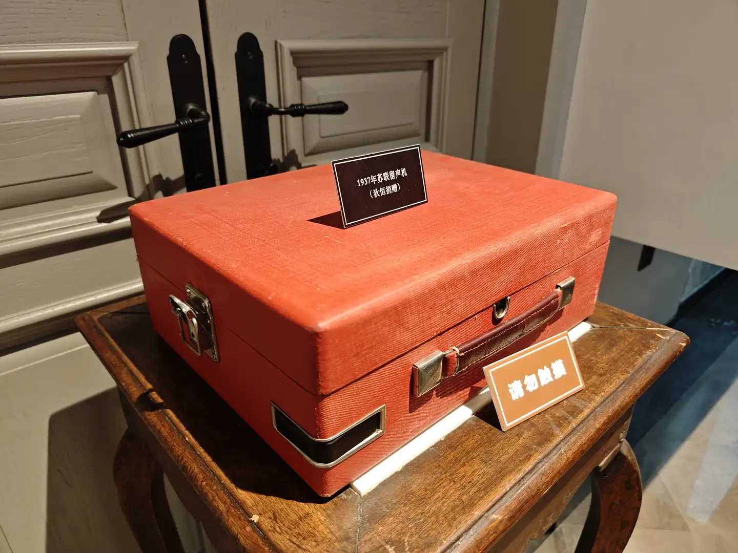 一个红色的方形盒子放在木质桌子上，盒子上方有一个黑色标签写着“1937年苏联留声机（冰恒捐赠）”，旁边还有一个提示牌写着“请勿触摸”