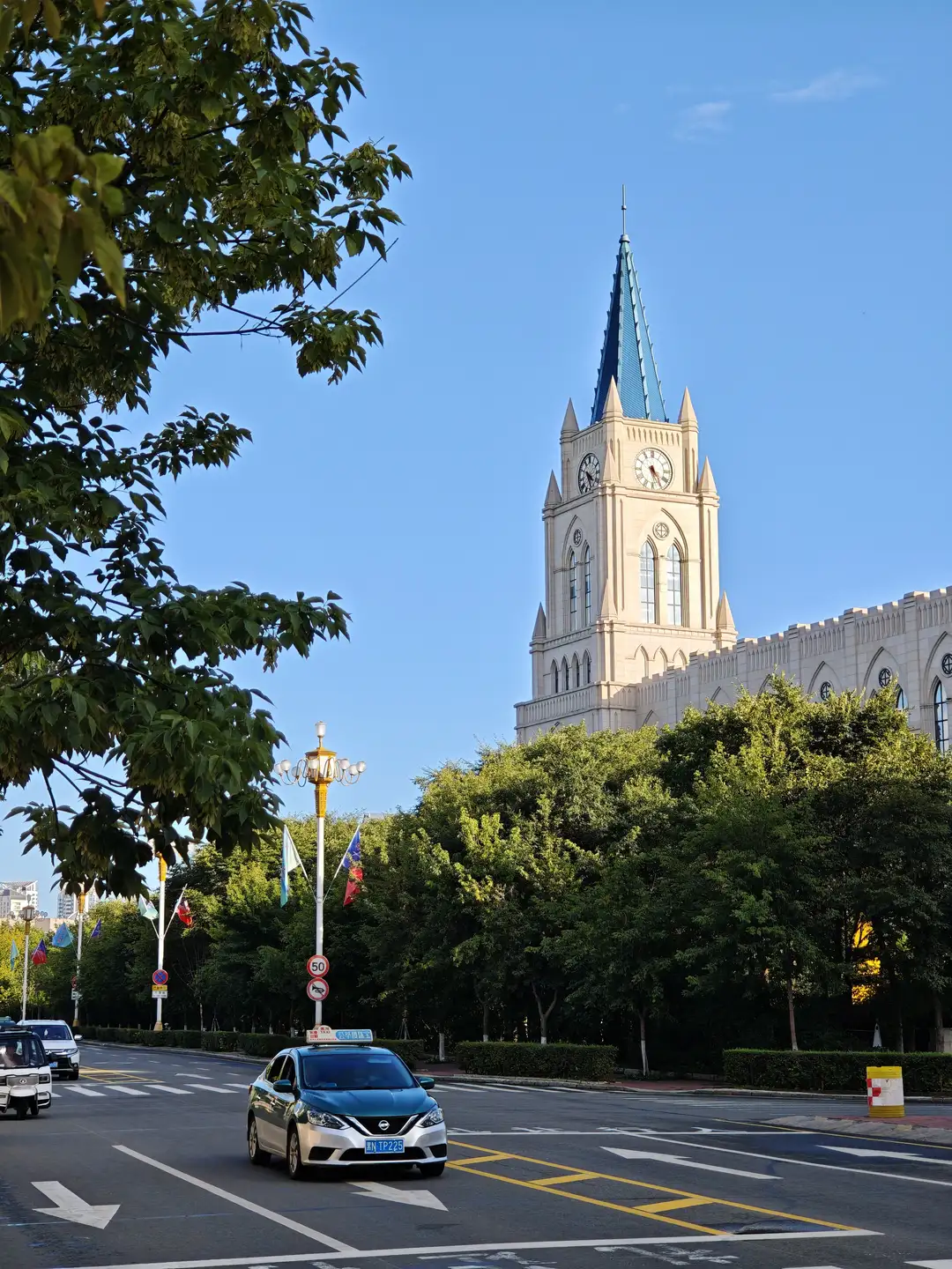 一座高耸的钟楼建筑位于街道尽头，塔顶为蓝色尖顶，塔身有多个拱形窗户和时钟，周围绿树成荫，道路上有行驶的车辆和路灯