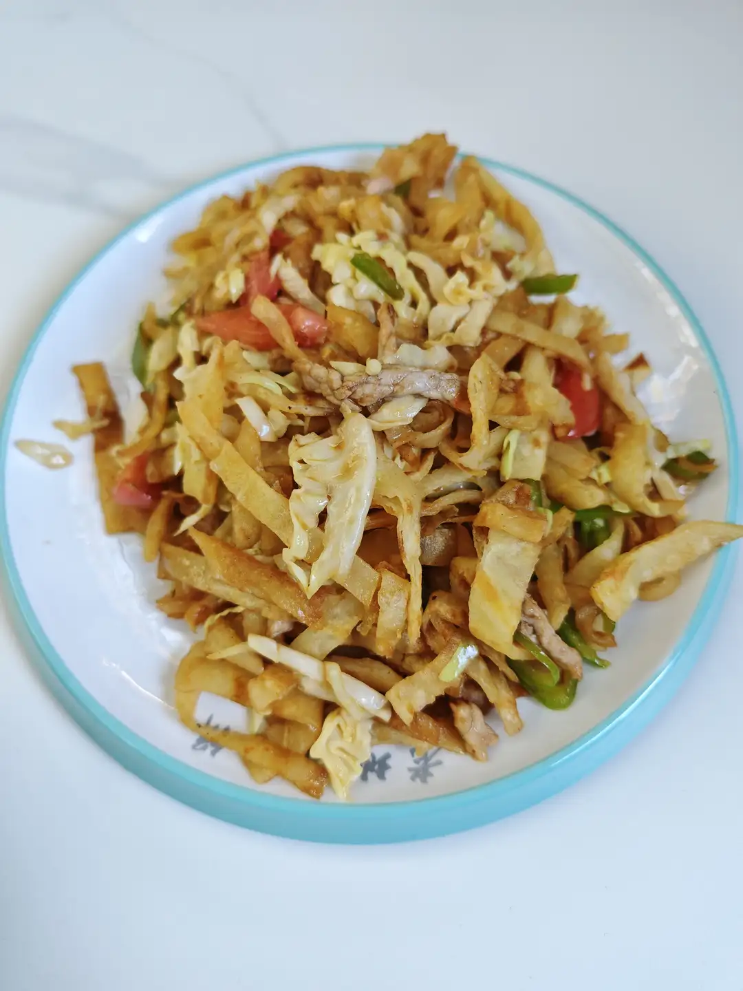 一盘炒菜盛在带有蓝色边框的白色盘子里，菜肴包含切片的土豆、卷心菜、青椒、红椒和肉丝，整体呈金黄色调