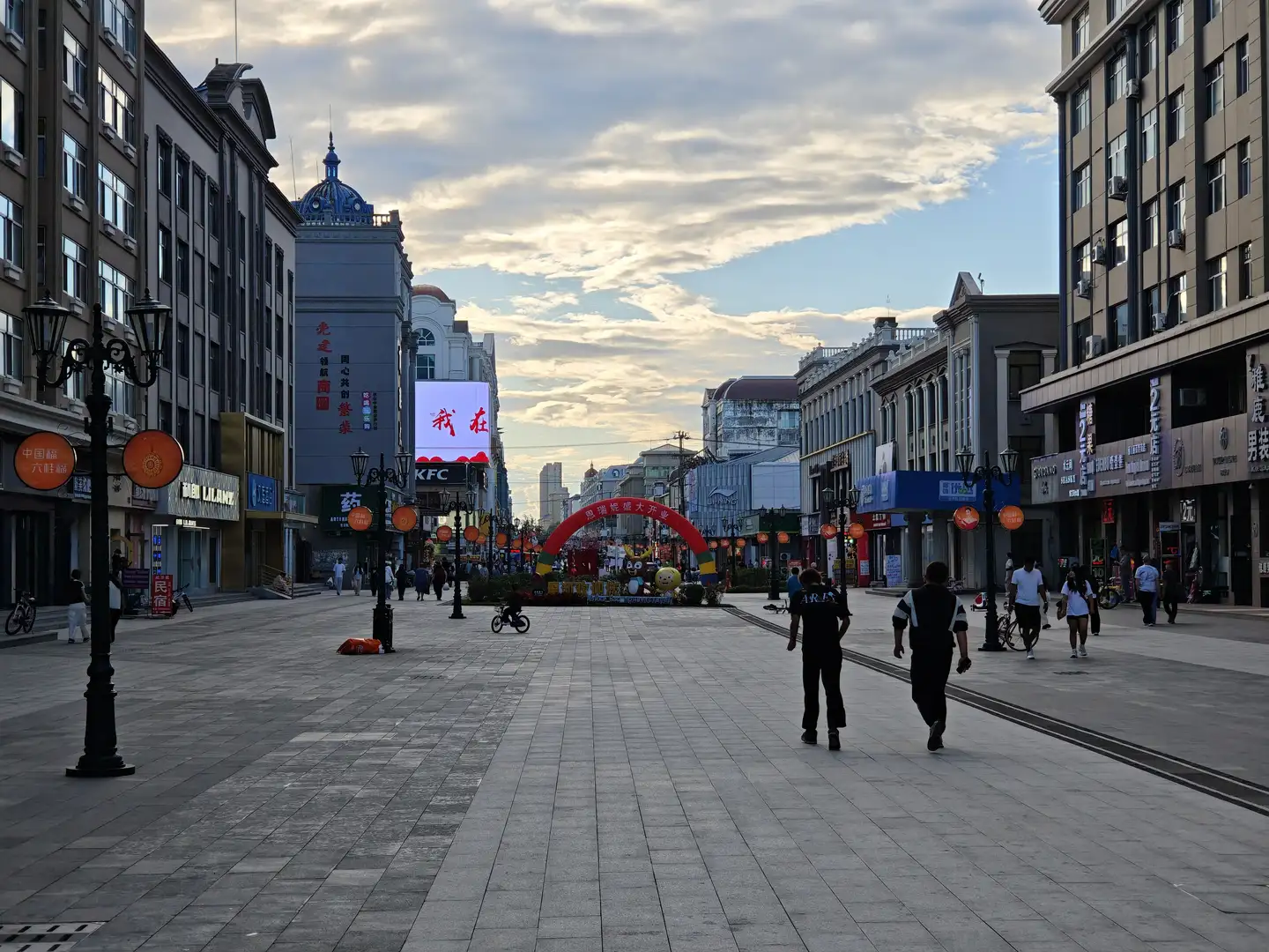 一条宽阔的步行街两旁是多层建筑，街道中央有装饰拱门和花坛，远处可见大型电子屏幕显示“我在”，行人和骑车人穿行其间