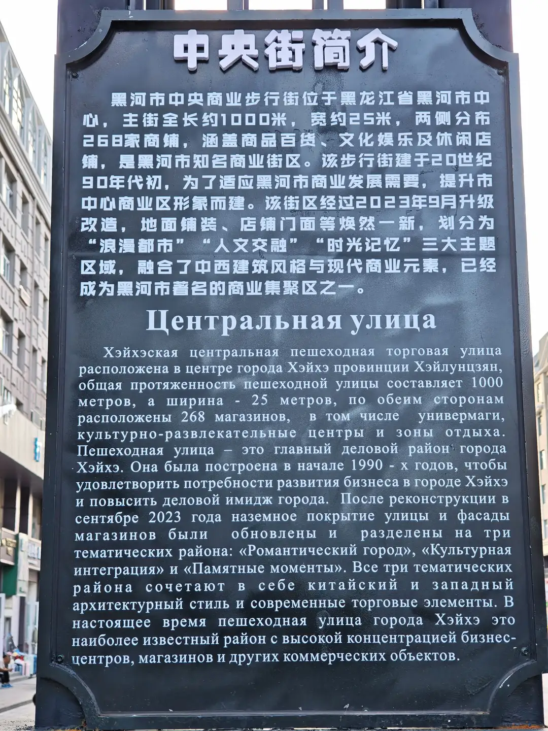 一块黑色的介绍牌立于街道旁，上面用中文和俄文介绍了黑河市中央商业步行街的历史、长度、宽度及改造情况，内容包括其主题分区和建筑风格融合等信息