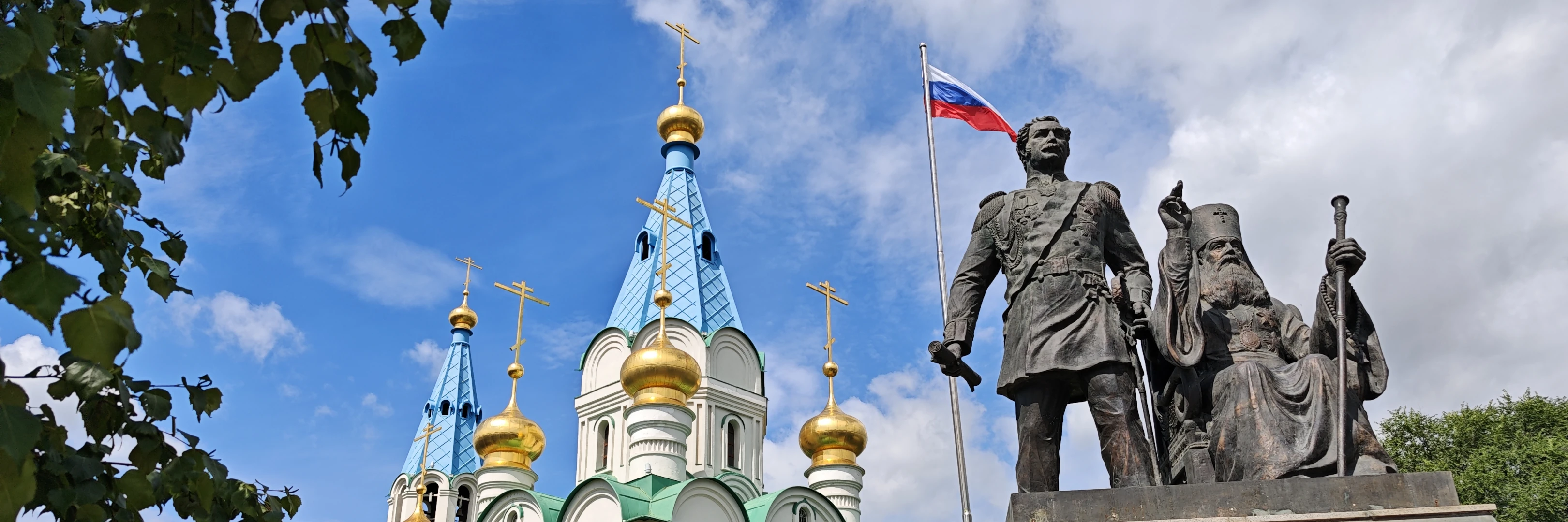 一座大型白色教堂，拥有多个蓝色和金色圆顶，前方矗立着一座雕像，雕像上有两名人物，一人站立持剑，一人坐着持权杖。教堂前有黑色铁艺围栏，俄罗斯国旗在风中飘扬。