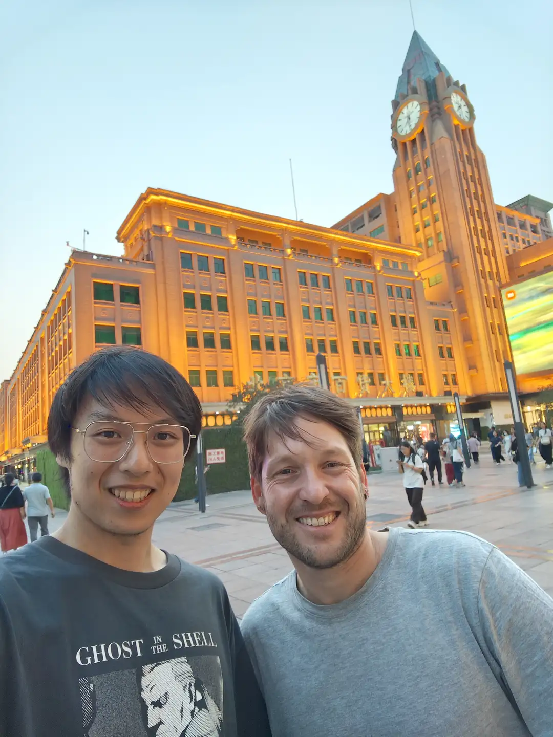 两个人站在一座大型建筑前，建筑上有明亮的灯光和一个高大的钟塔。建筑的外墙呈黄色，窗户排列整齐。背景中有一些行人和绿色植物。