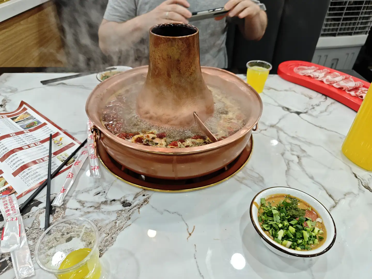 桌子上有一个铜制火锅，锅内有热气升起，旁边放着一碗蘸料和一些餐具。火锅分为两个部分，一侧有红色辣椒。背景中有人正在用手机拍照。