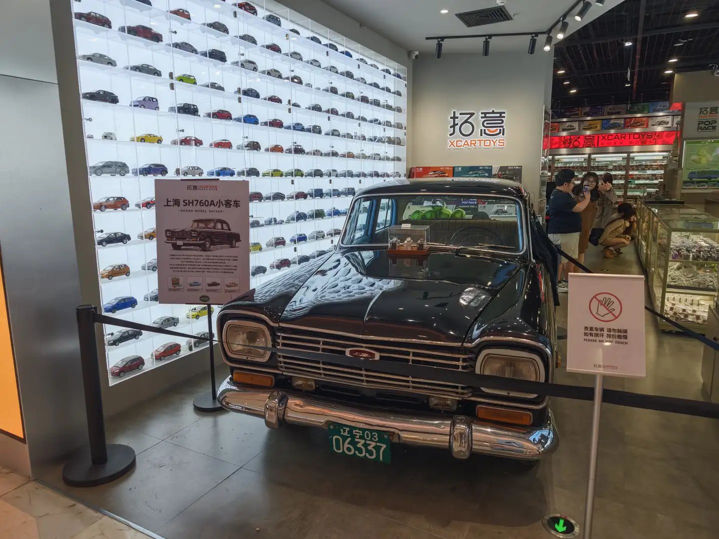 展厅内展示了一辆黑色复古汽车，车牌号为辽宁03 06337，旁边有一个标牌介绍汽车信息。墙上陈列着许多小型汽车模型，形成一面汽车墙。展厅内还有几位参观者在观看展品。