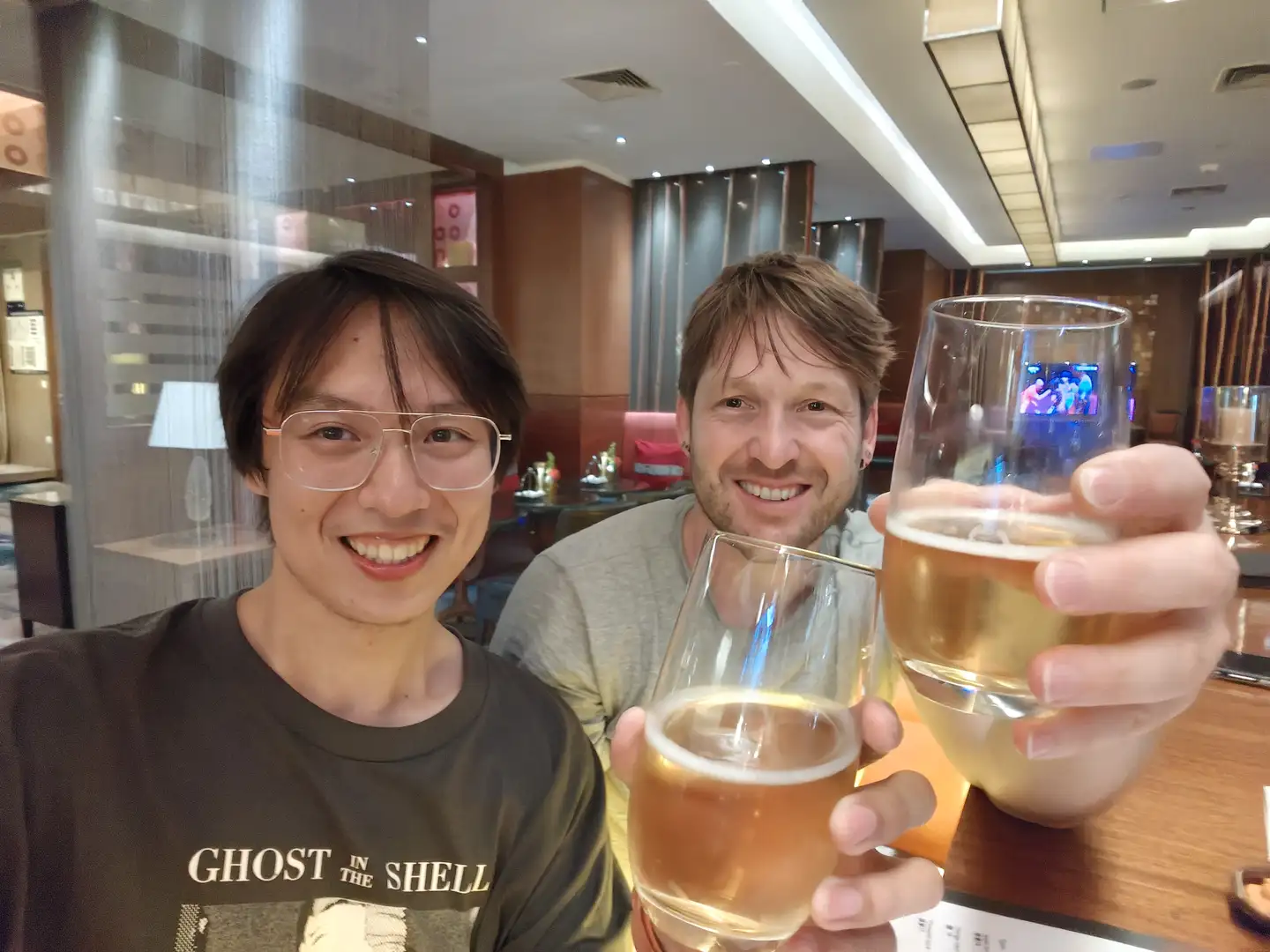 两个人举着啤酒杯，杯子中有浅色液体，背景是一个室内环境，装饰现代。他们穿着休闲服装，一人穿灰色T恤，另一人穿印有“GHOST IN THE SHELL”字样的黑色T恤。背景中可以看到一些餐厅设施和装饰。