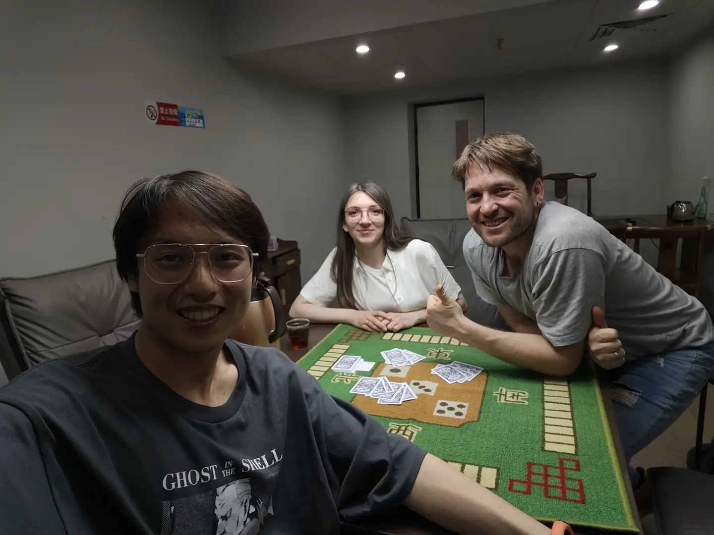 三个人坐在桌子旁，桌上铺着绿色麻将桌布，上面摆放着麻将牌。左边的人穿着印有“GHOST IN THE SHELL”字样的黑色T恤，右边的人穿着灰色T恤，中间的人穿着白色上衣。背景是一个室内房间，墙上贴有禁止吸烟标志。