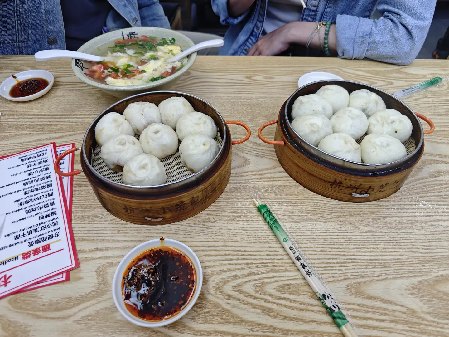 桌上摆放着两笼包子、一碗汤和一些调料。旁边有一双筷子和菜单，菜单上列有各种面食选项。桌子是木质的，表面有纹理。