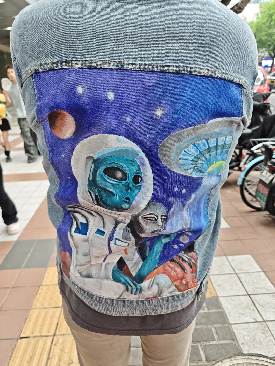 一个人穿着牛仔夹克，夹克背面绘有太空人和外星人的图案。背景是街道场景，地面铺有砖块。旁边有行人和自行车。