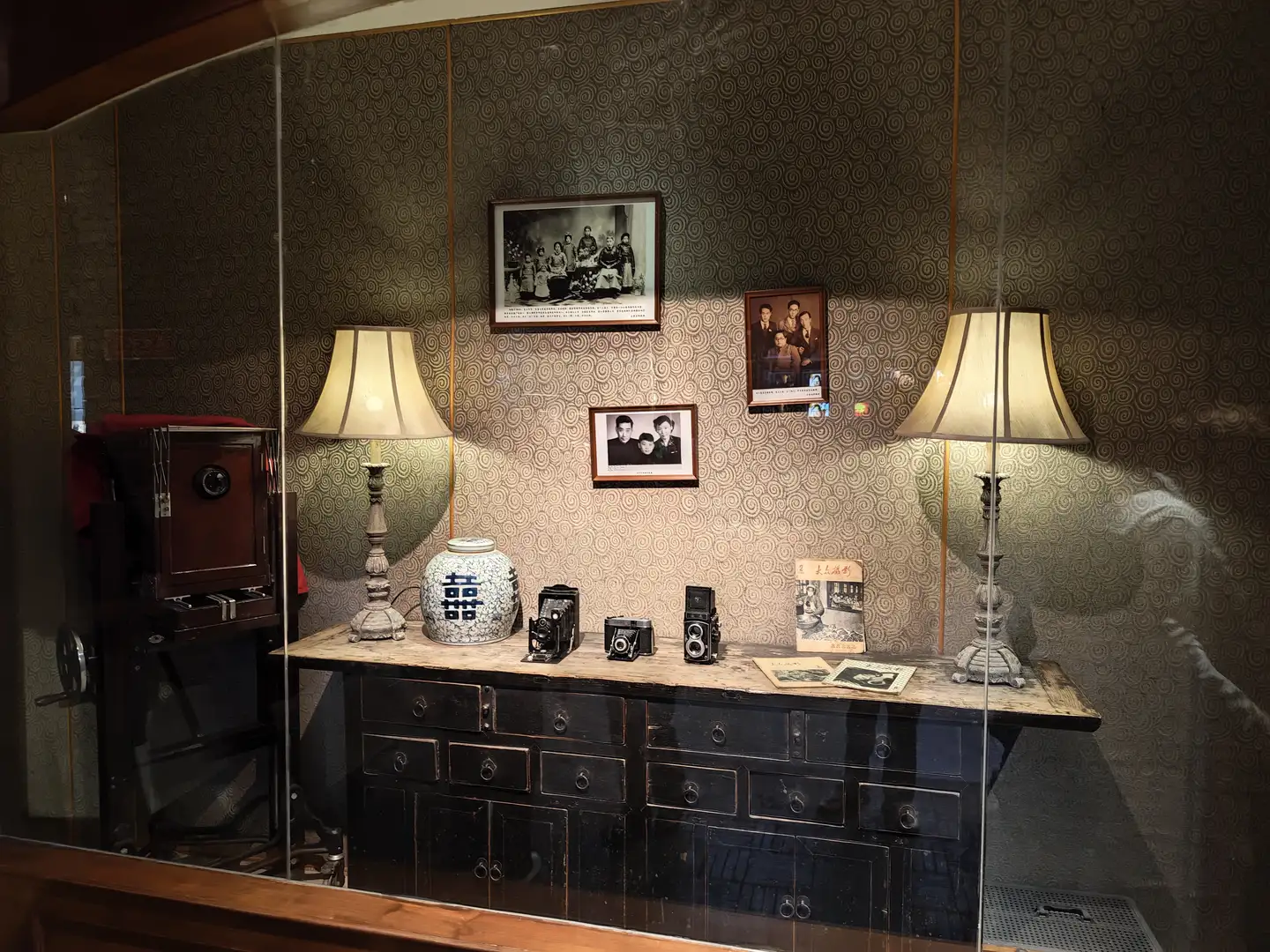 玻璃柜内展示了一张旧式照片墙，墙上挂着几张黑白照片。柜子上摆放着几台老式相机和一个带有双喜字样的瓷罐。柜子两侧各有一盏台灯。
