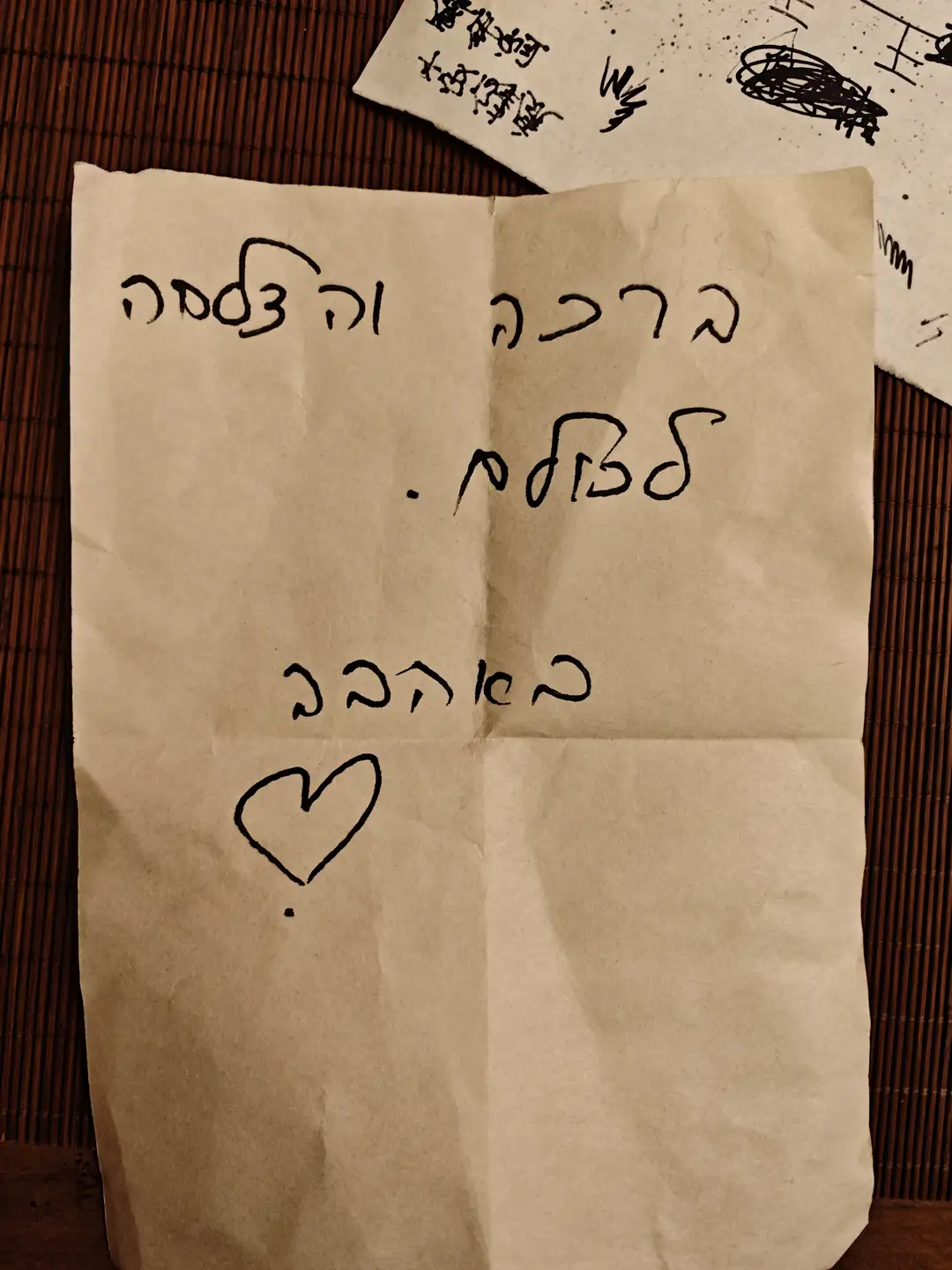 一张纸上用希伯来文写着内容，纸上有折痕和一个心形符号。纸张放在竹席上，旁边还有另一张部分可见的纸。文字清晰可见。