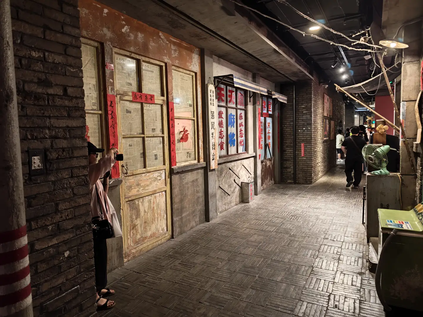 一条走廊两侧是砖墙结构，墙上贴有报纸和春联。走廊地面铺有方形瓷砖，远处有人行走。左侧一人正在拍照。