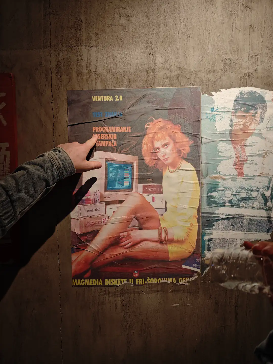 墙上贴着一张复古海报，海报上有一位金发女性坐在电脑前。海报上方写着“VENTURA 2.0”，下方有“MAGMEDIA DISKETE U FRI-ŠOPOVIMA”字样。一只手正指着海报。