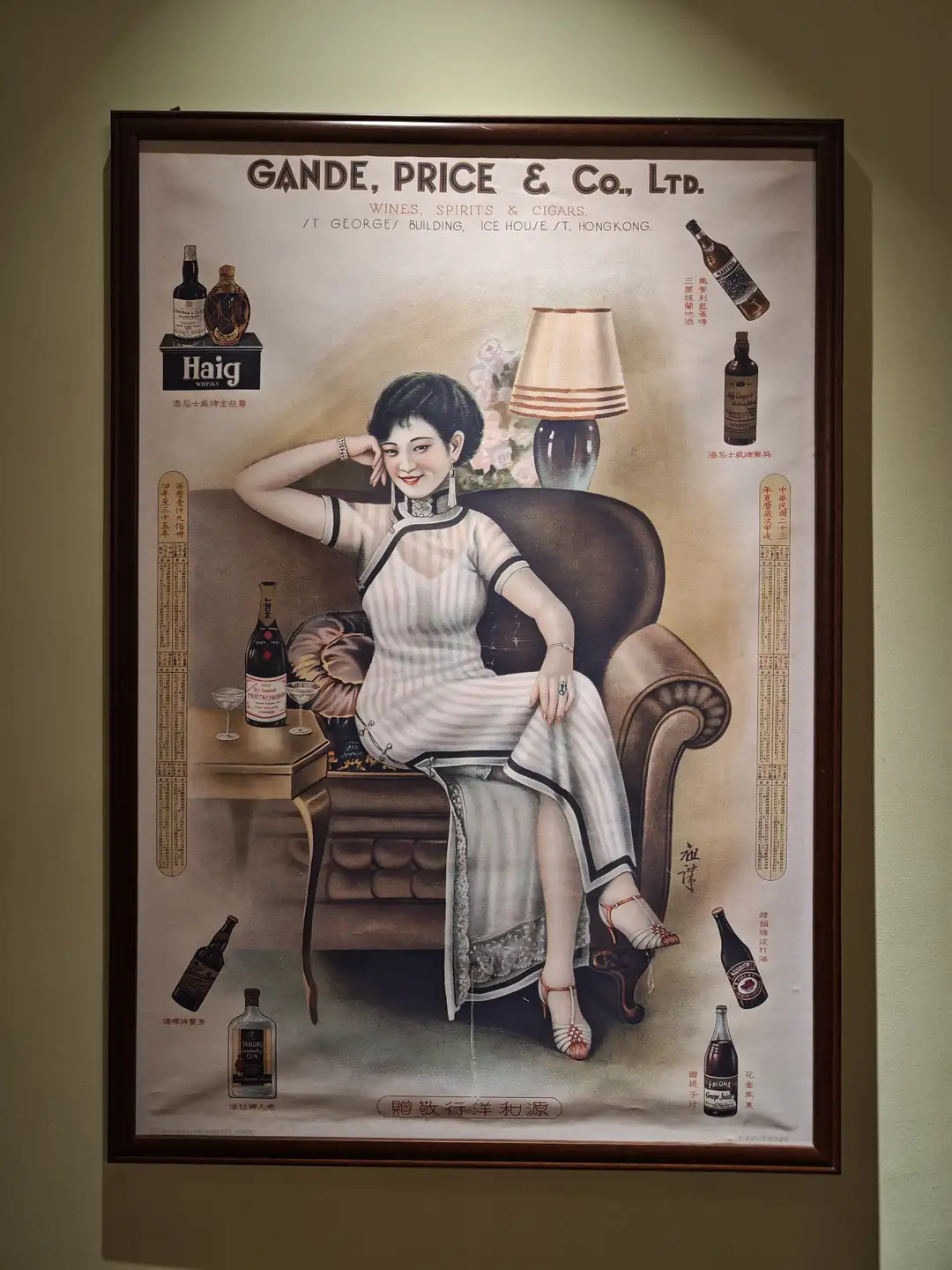 一幅复古广告画中，一位女性坐在沙发上，周围摆放着酒瓶和酒杯。广告标题为“GANDE, PRICE & Co., LTD.”，并提到“WINES, SPIRITS & CIGARS”。画框为深色木制。