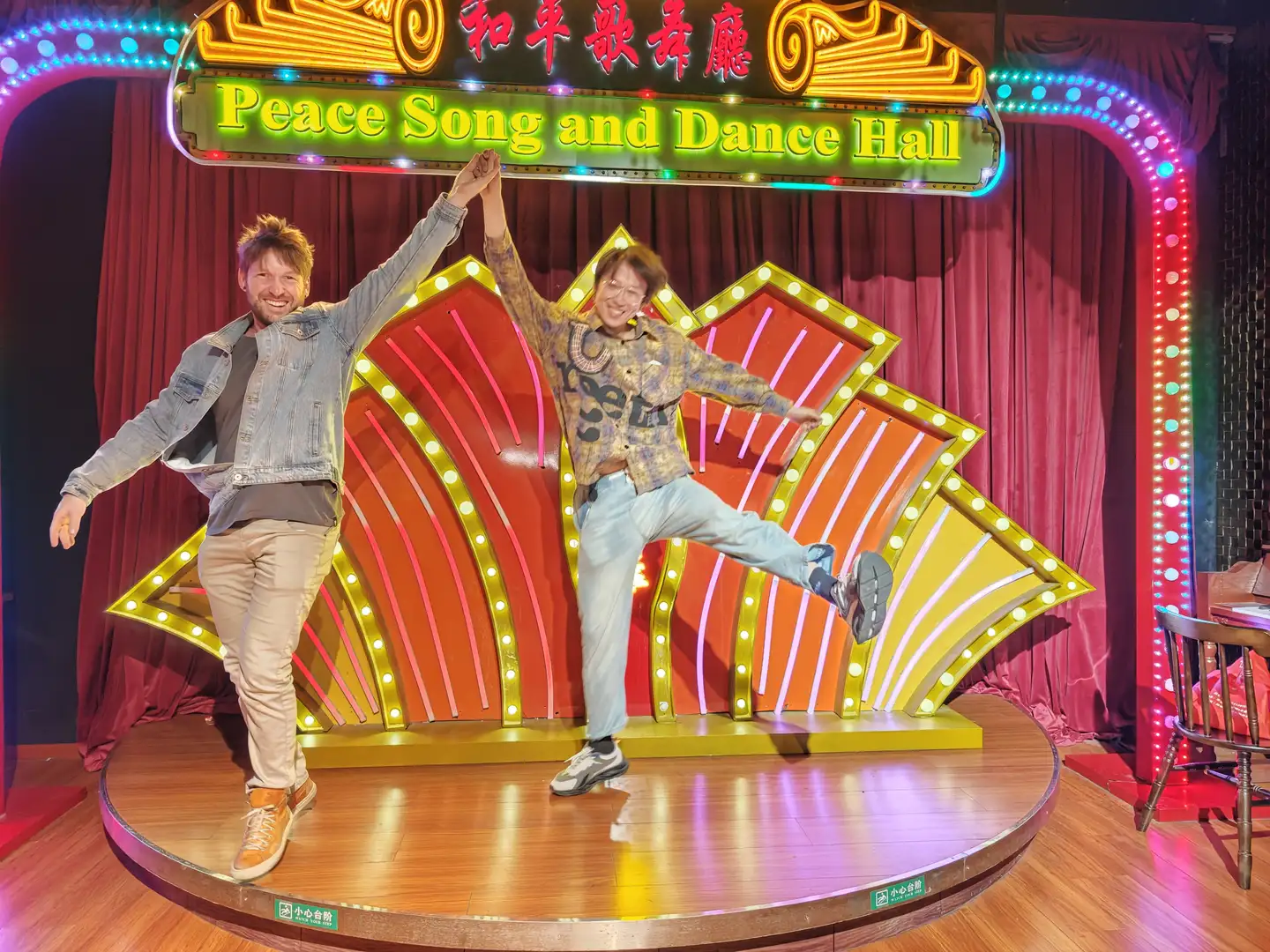 两个人站在舞台上，舞台背景标有“Peace Song and Dance Hall”的霓虹灯牌。左边的人穿着浅色衣服，右边的人单腿抬起，穿着蓝色裤子。舞台灯光闪烁。