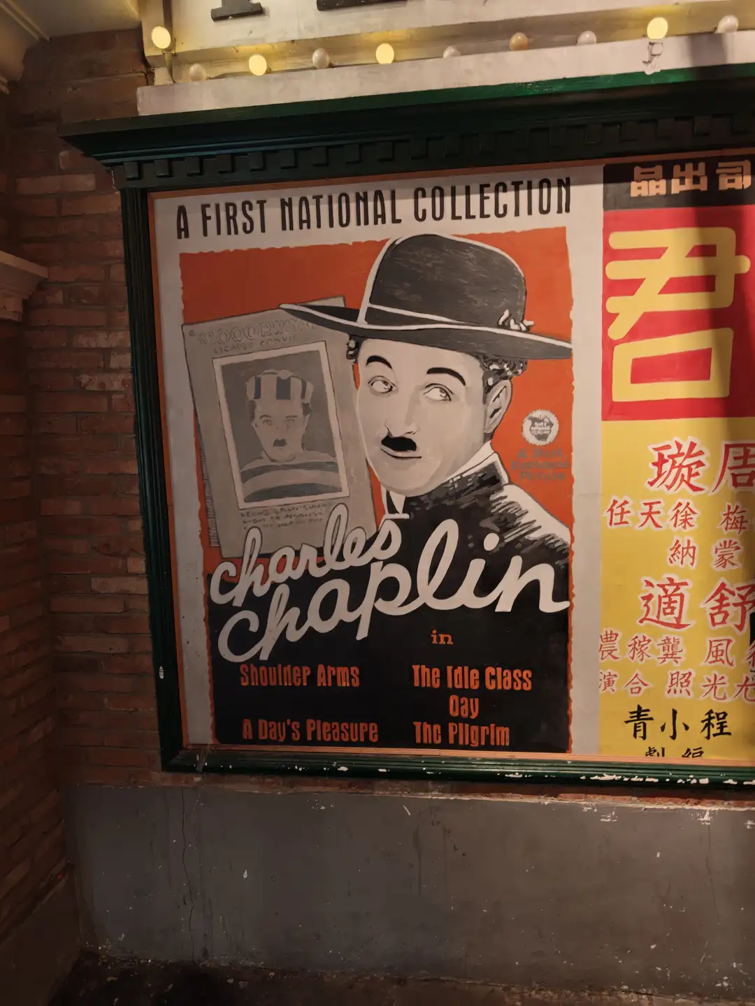 墙上挂着一张查理·卓别林的电影海报，海报上写有“A FIRST NATIONAL COLLECTION”和多部电影名称。海报右侧有中文文字，背景为红砖墙。海报上方有灯光装饰。