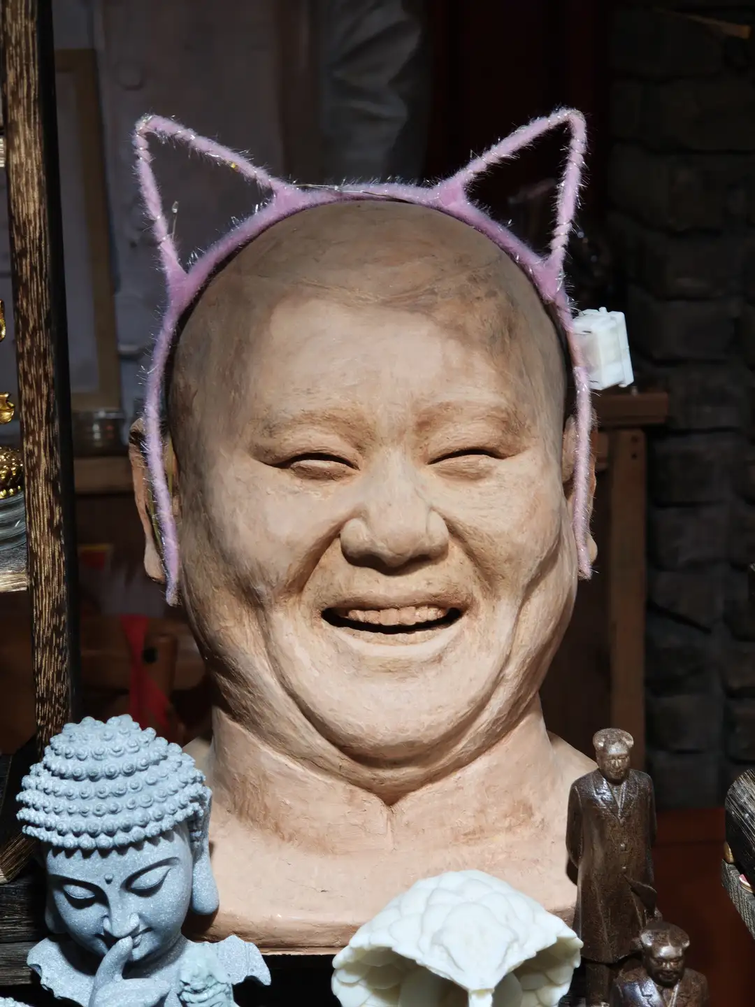 一个雕塑头像戴着粉色猫耳发箍，旁边有佛像和人物雕像。雕塑表情微笑，眼睛微闭。背景为室内环境，有木质架子和其他物品。