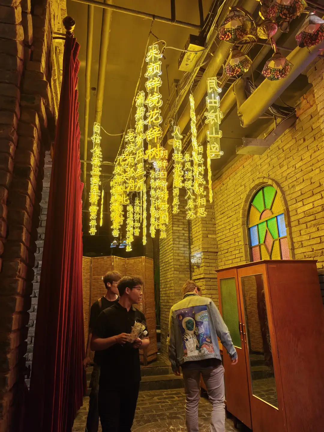 一条走廊装饰着黄色霓虹灯，墙上挂着中文诗句。几个人在走廊中行走，其中一人穿着印有宇航员图案的夹克。走廊尽头有彩色玻璃窗。