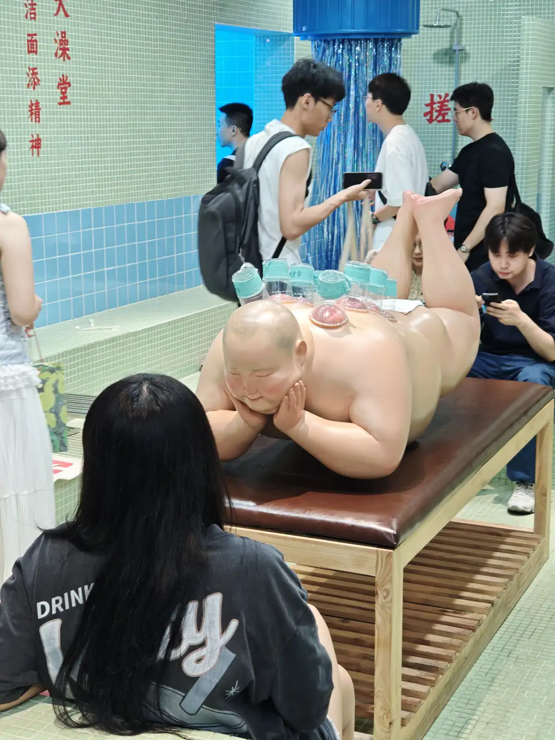 一个大型人体模型躺在长椅上，周围有多人观看或拍照。模型姿态放松，头部枕在手臂上。背景为浴室环境，墙壁上有蓝色瓷砖。