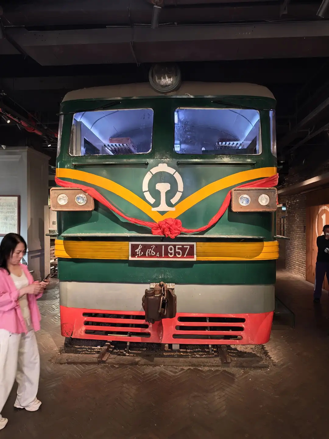 一辆绿色火车头停在室内展览空间，车头前系着红色绸带。火车编号为东风4 1957，前方有圆形标志。一名女子站在火车旁看手机。