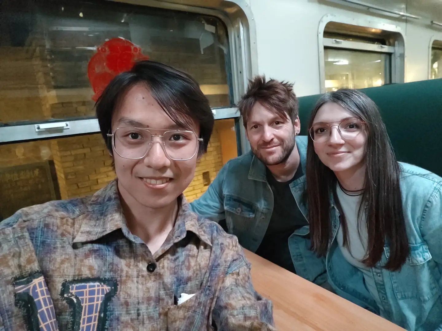 三个人坐在桌子旁合影，中间的人穿着牛仔外套，背后是砖墙和窗户。左边的人戴眼镜，穿格子衬衫。右边的人也戴眼镜，穿浅色衣服。